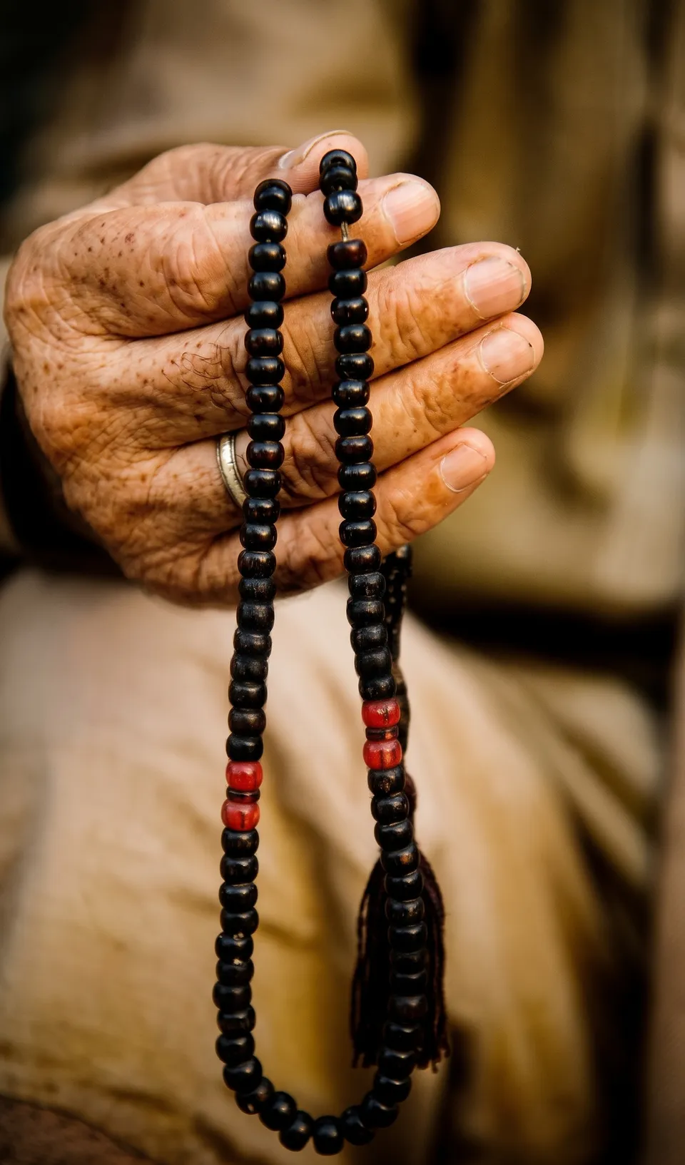 An elderly hand holding black prayer beads, with a few red beads, in warm light.